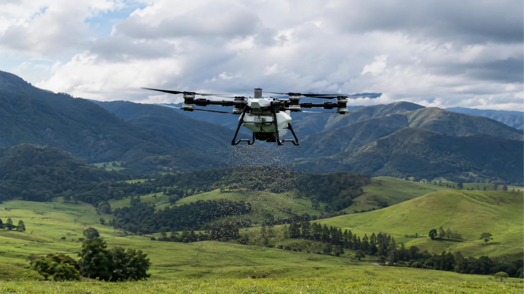 A drone spreading granular fertiliser and seed over green hill country