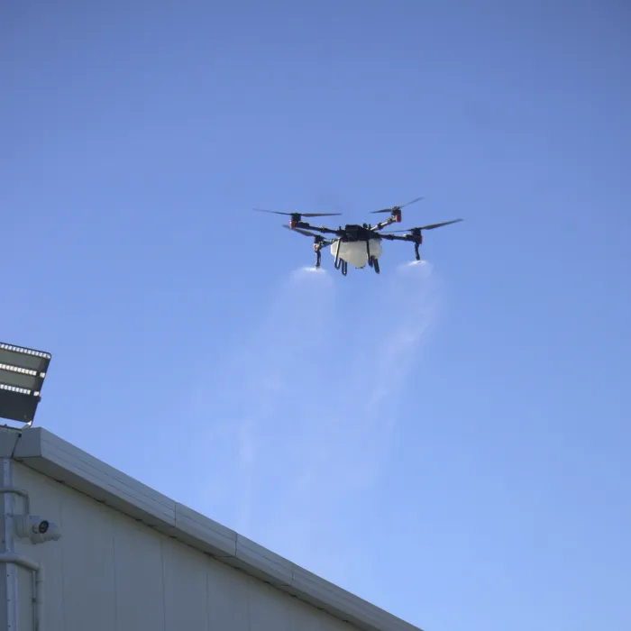 A drone applying moss and mould treatment above a commercial shed roof against a blue sky